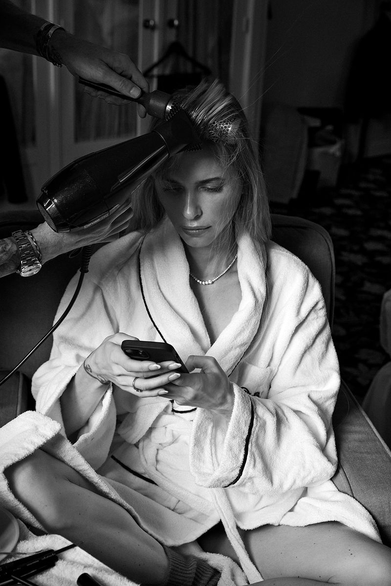 A woman in a white robe sits while someone styles her hair with a blow dryer. She's looking at her cell phone indoors.