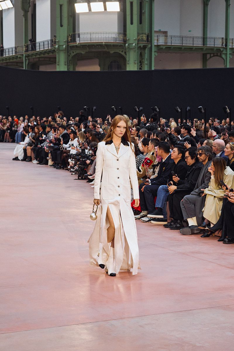 Runway model in white coat-dress walks during a fashion show, front view, surrounded by an audience and photographers inside an event space.