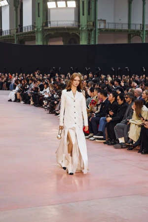 Runway model in white coat-dress walks during a fashion show, front view, surrounded by an audience and photographers inside an event space.
