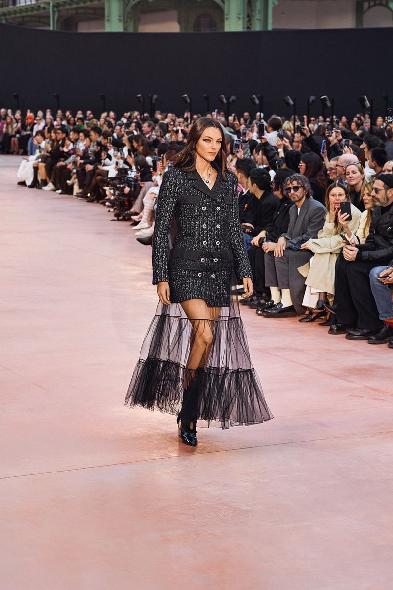 Runway model showcases a black tweed dress with a layered tulle skirt during a fashion show, with a blurred audience in the background.