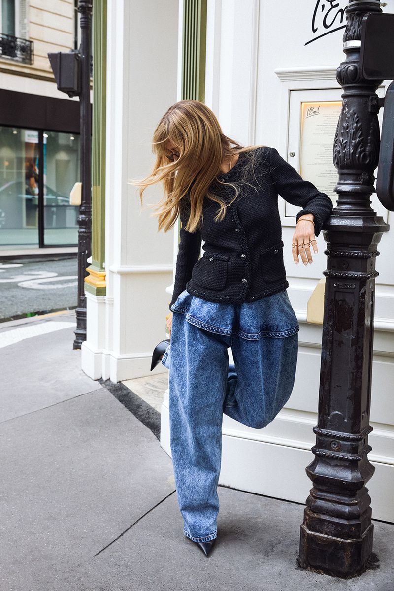 Blonde woman in a black tweed jacket and acid-wash jeans posing on a city street next to a vintage-style street light.