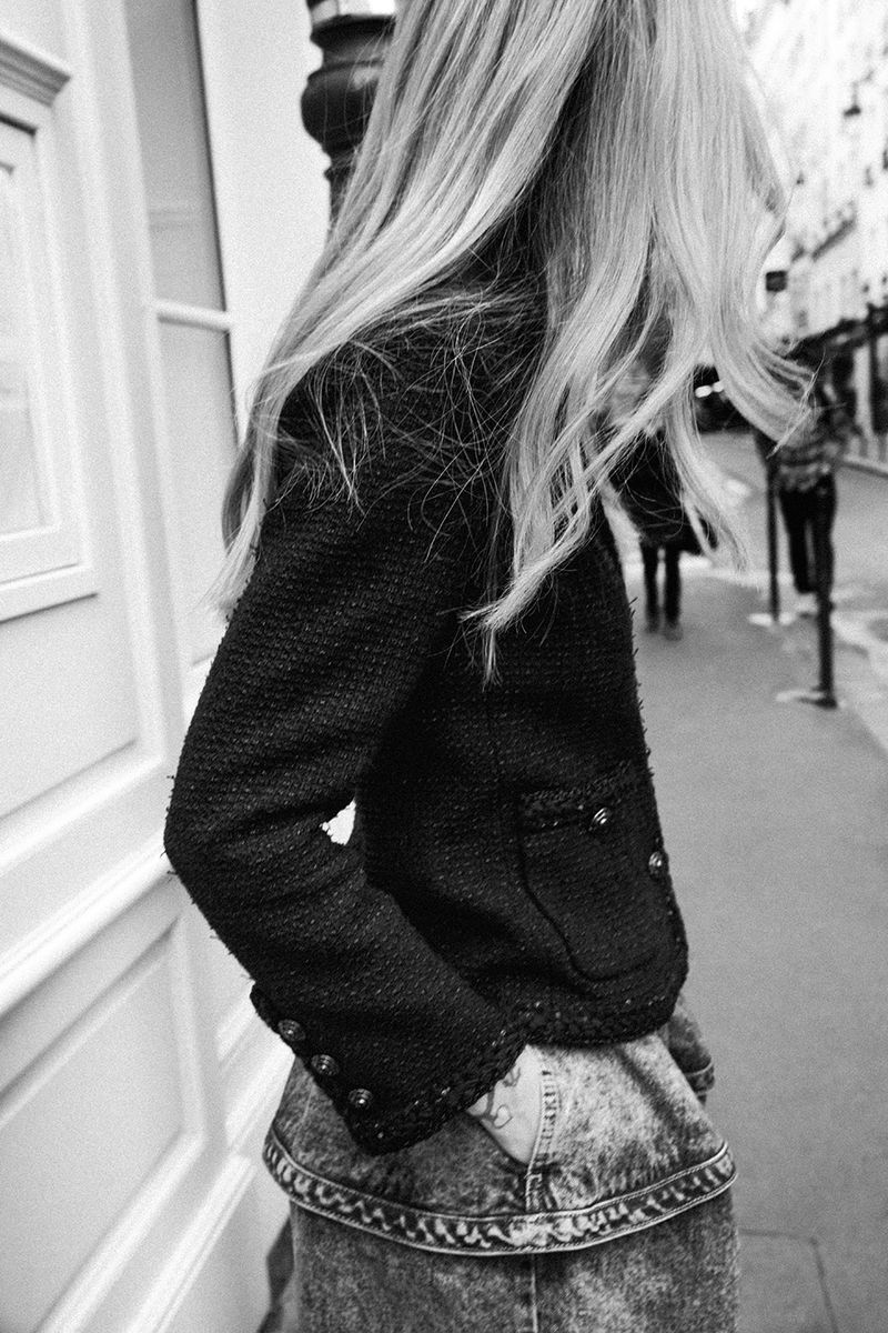 Woman in jacket standing on city street, looking away from the camera in black and white. Fashion editorial street-style photo.