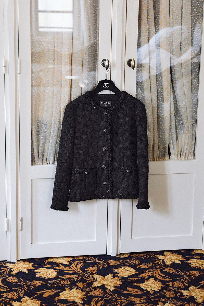Black Chanel jacket hanging on a hanger in front of paneled glass doors in a home interior with a dark floral carpet.