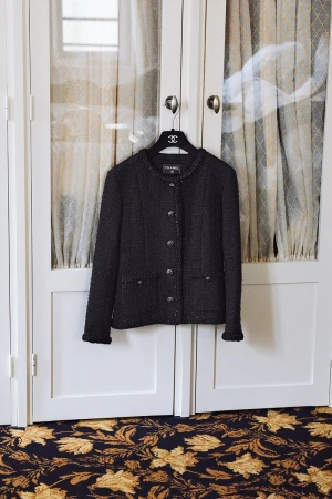 Black Chanel jacket hanging on a hanger in front of paneled glass doors in a home interior with a dark floral carpet.