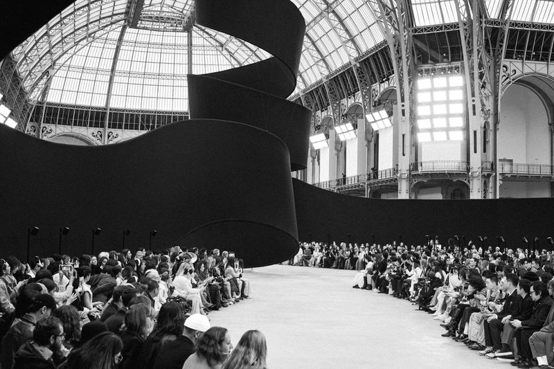 Fashion show audience seated in a grand hall with a spiral backdrop; focus on crowd and interior architecture in black and white.