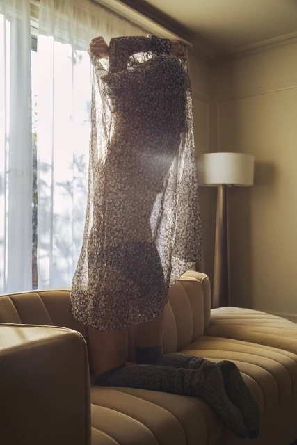 Woman kneeling on a gold couch in a floral dress and sheer robe in a warm-toned interior with natural window light and a table lamp.