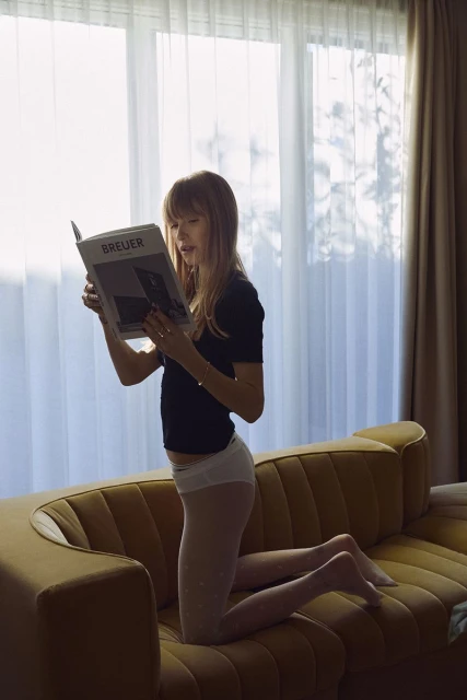 Woman on knees reading book titled 'BREUER' near bright window, wearing black shirt, light tights, lounging on yellow couch. Indoors.