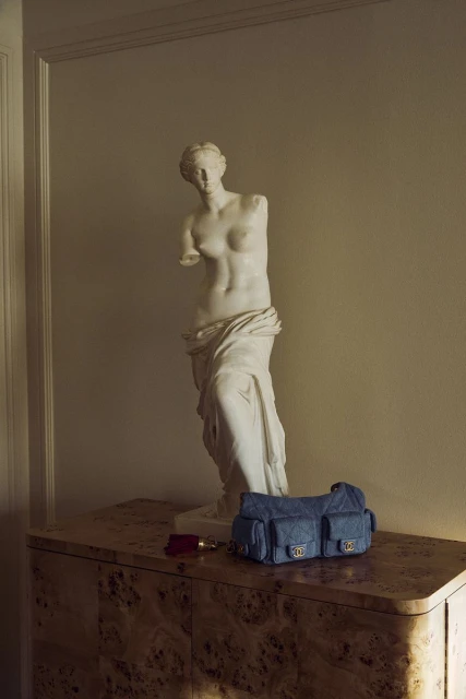 Statue of a woman with a blue Chanel handbag and red wallet on a wooden cabinet in an indoor setting with neutral wall paint.
