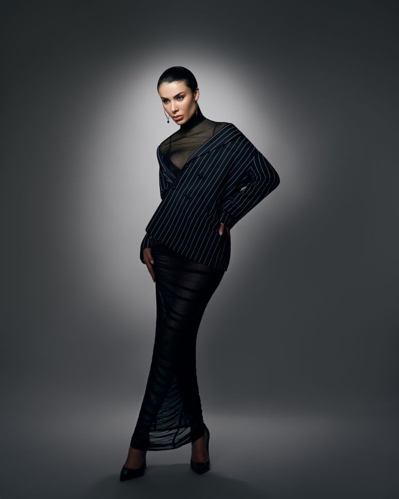 Woman poses in pinstripe blazer and black dress in a studio with gray background and spot lighting, showcasing fashion and beauty.