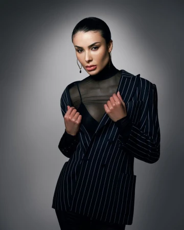 Woman in pinstripe blazer and black top poses confidently in a studio with soft, directional lighting and a neutral grey background.