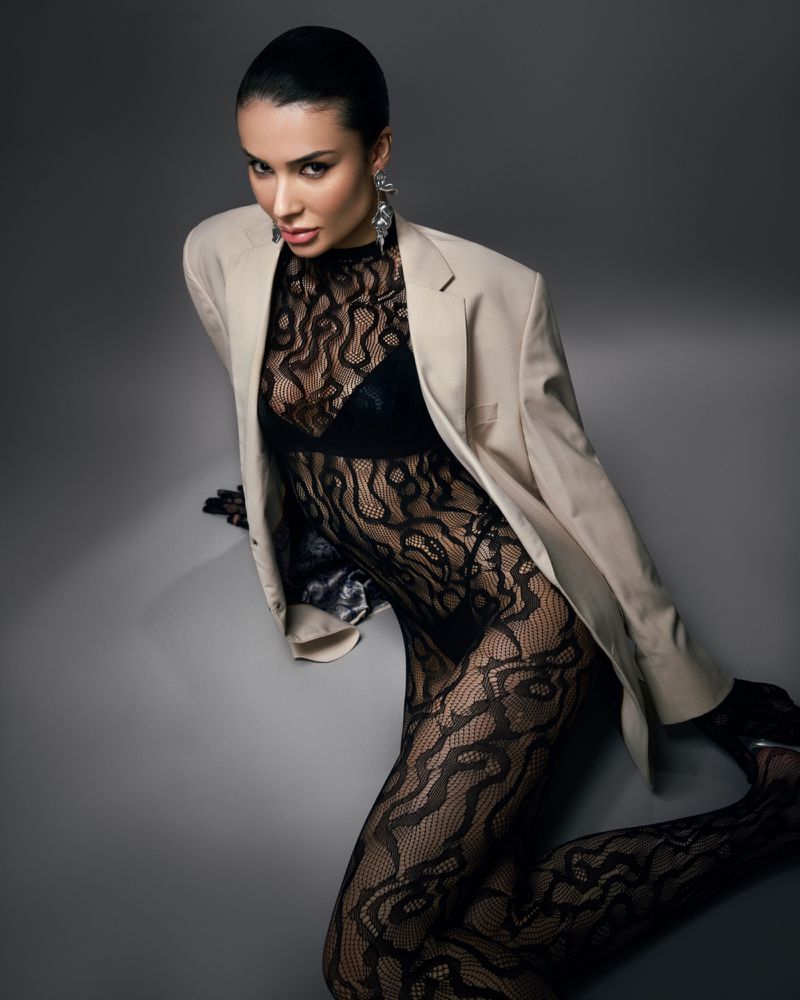 Portrait of a woman with dark hair, wearing a nude-colored blazer, black lace top and leggings, and large earrings, posing against a gray background.