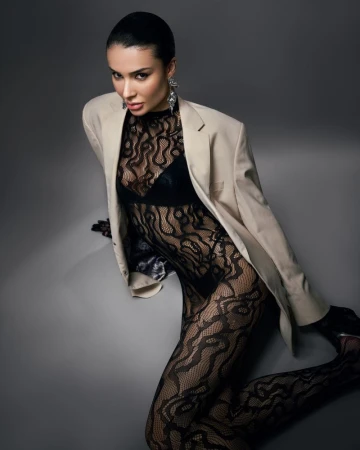 Portrait of a woman with dark hair, wearing a nude-colored blazer, black lace top and leggings, and large earrings, posing against a gray background.