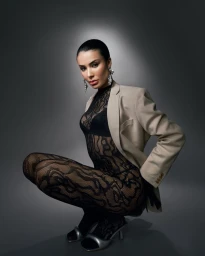Woman squatting in a black lace bodysuit, beige blazer, and silver heeled sandals against a dark grey gradient studio background