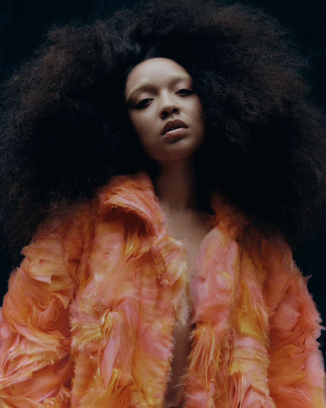 Portrait of a woman with afro hairstyle wearing a orange faux fur coat against a dark background in studio lighting.