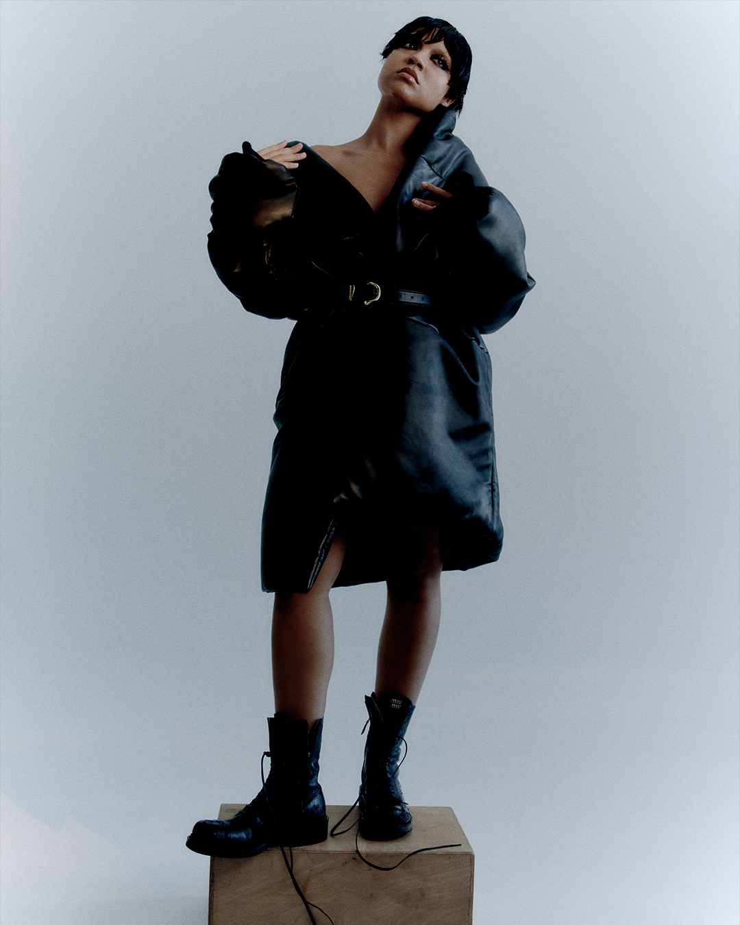 Fashionable young adult woman posing in oversized black leather coat and chunky black boots, standing on wooden block against grey background.