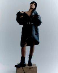 Model in black leather coat and combat boots standing on a wooden block against a plain grey studio backdrop