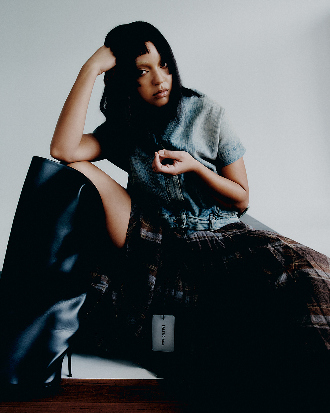Fashion shot of a dark-haired woman in denim top, plaid skirt and black boots, seated indoors against a grey wall in a studio setting.