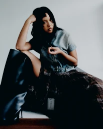 Model with dark hair wearing a denim top and plaid skirt with high black boots against a neutral background.