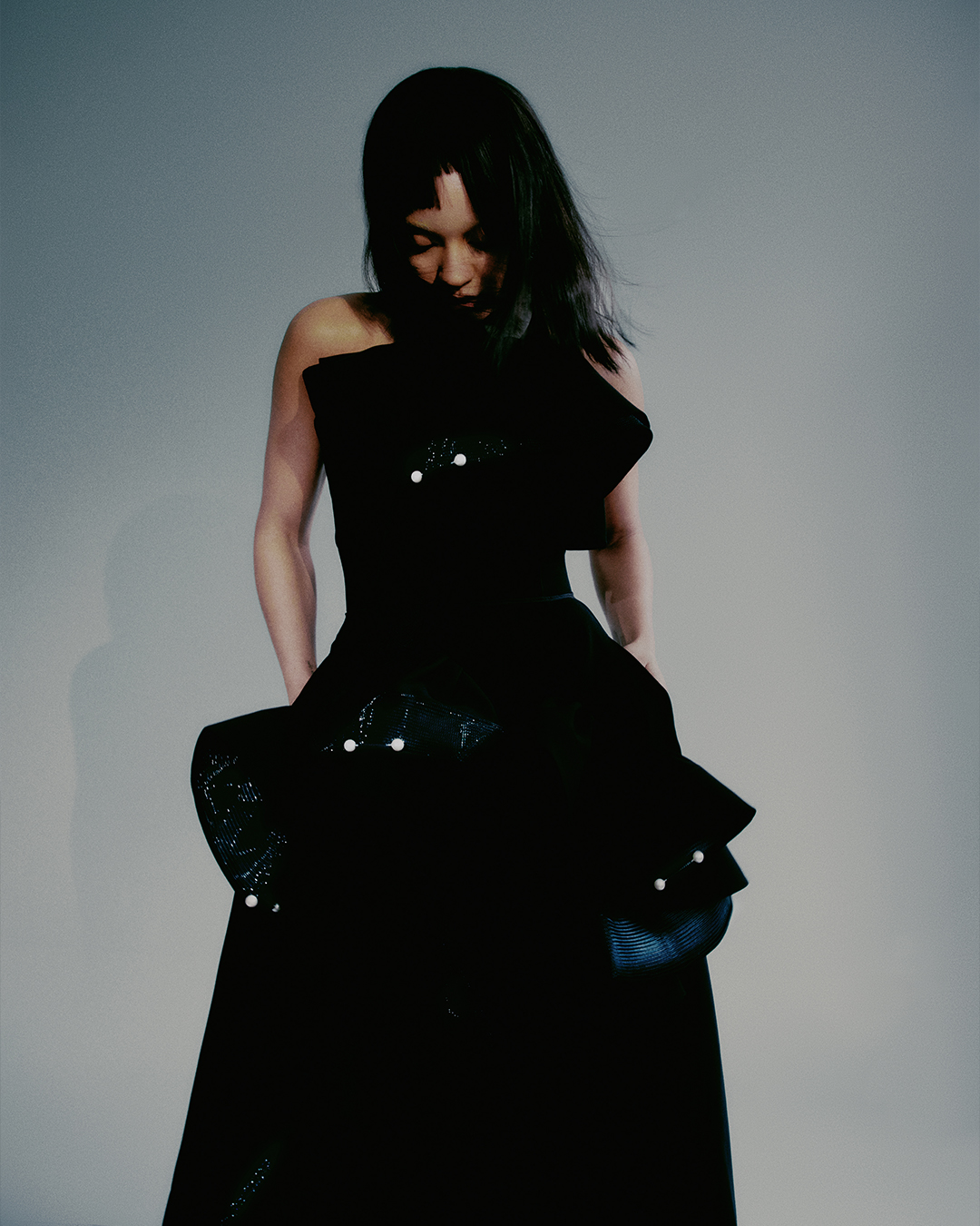 Woman in an elegant black strapless dress with embellishments, posing in front of a light grey backdrop in a fashion forward studio shot.