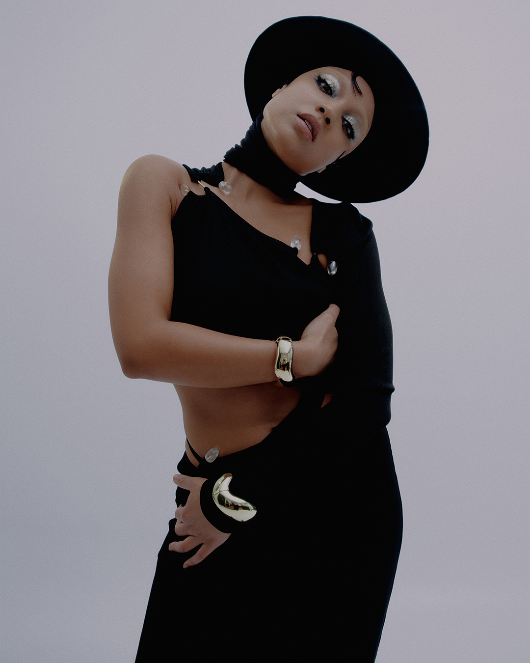 Portrait of a woman wearing a black hat and a black one-shoulder outfit with embellishments posing against a light grey background.