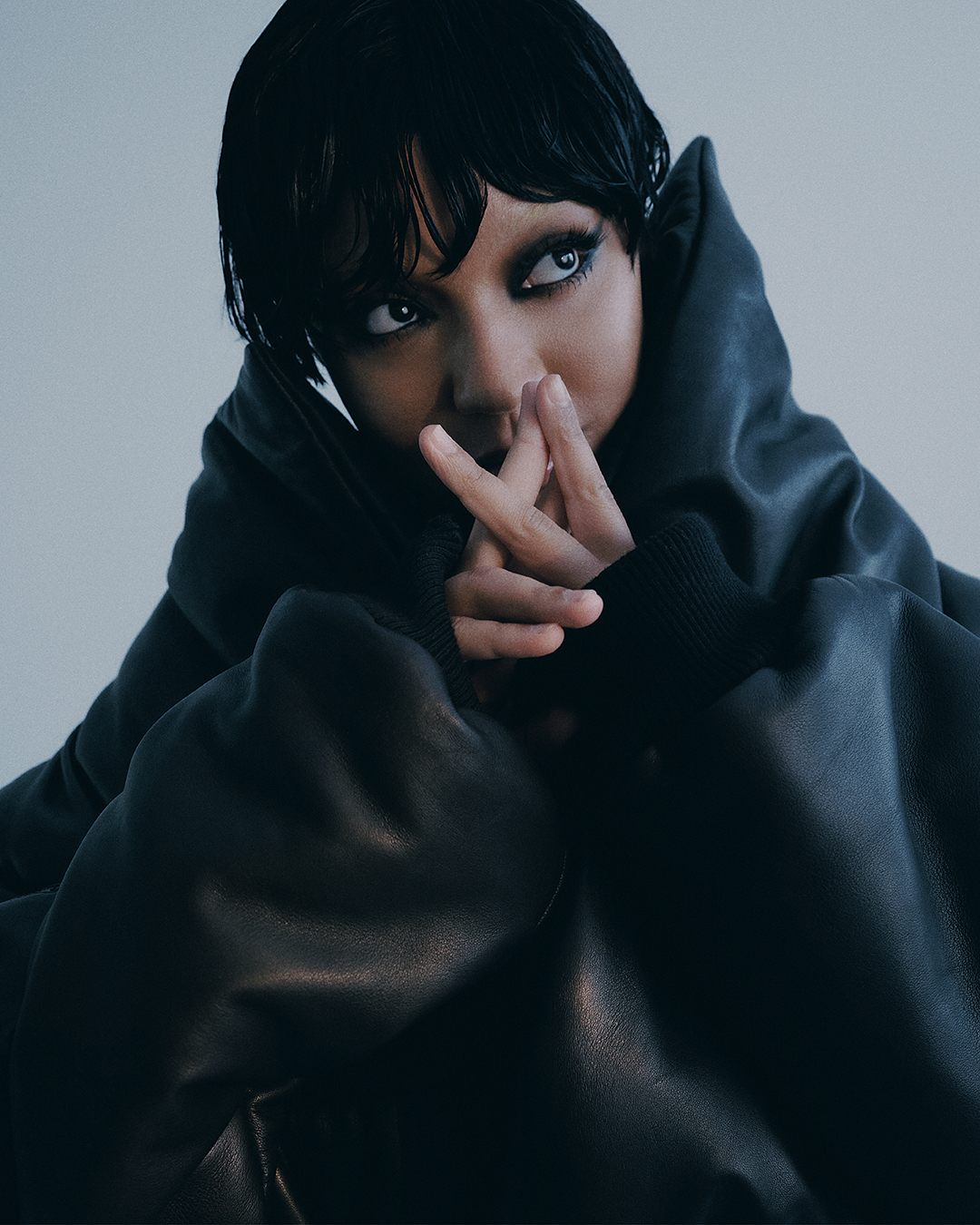 Dark-haired model with dramatic eye makeup and oversized hooded jacket poses indoors against neutral backdrop in close-up portrait.