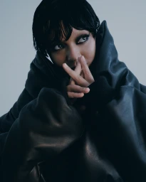 Model in a black puffer coat with dark eye makeup and wet-look black hair posing against a neutral grey studio background