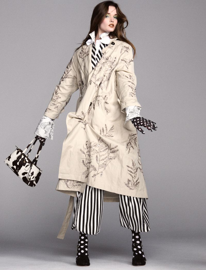 A woman models a long beige coat with dark leaf designs, striped pants, and polka-dot accessories against a grey studio backdrop.