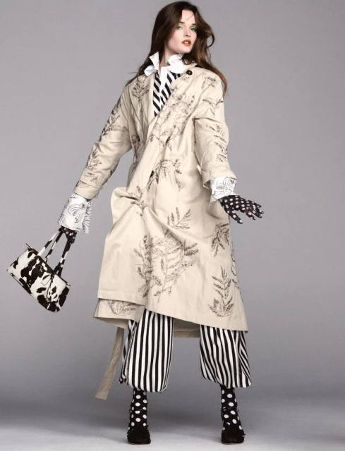 A woman models a long beige coat with dark leaf designs, striped pants, and polka-dot accessories against a grey studio backdrop.