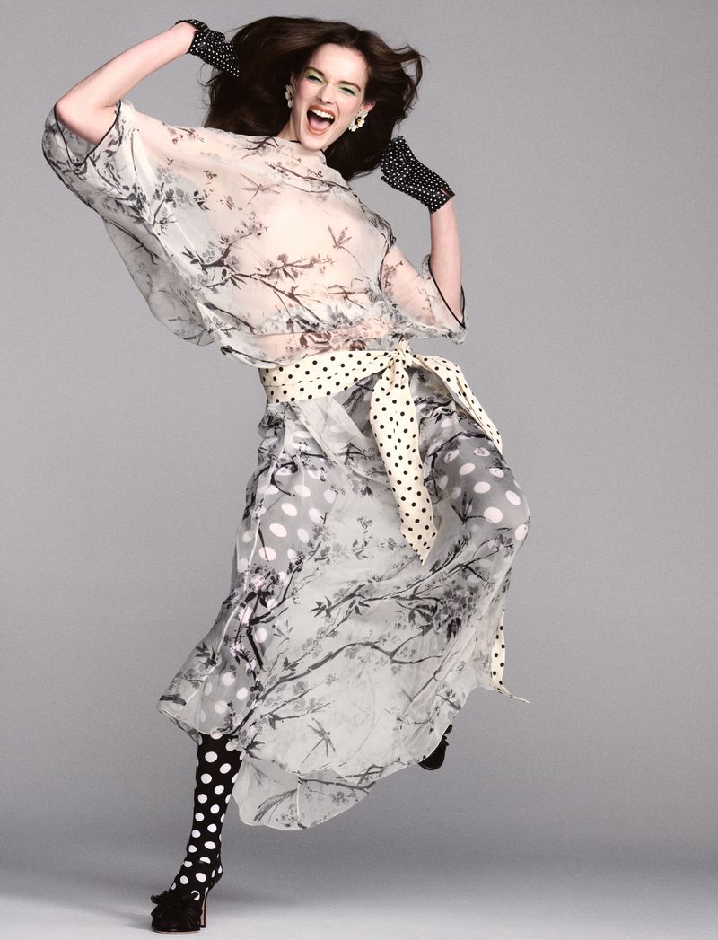 A woman in sheer, flowing, patterned separates poses expressively with arms up, black and white gloves, patterned stockings and heels on grey.