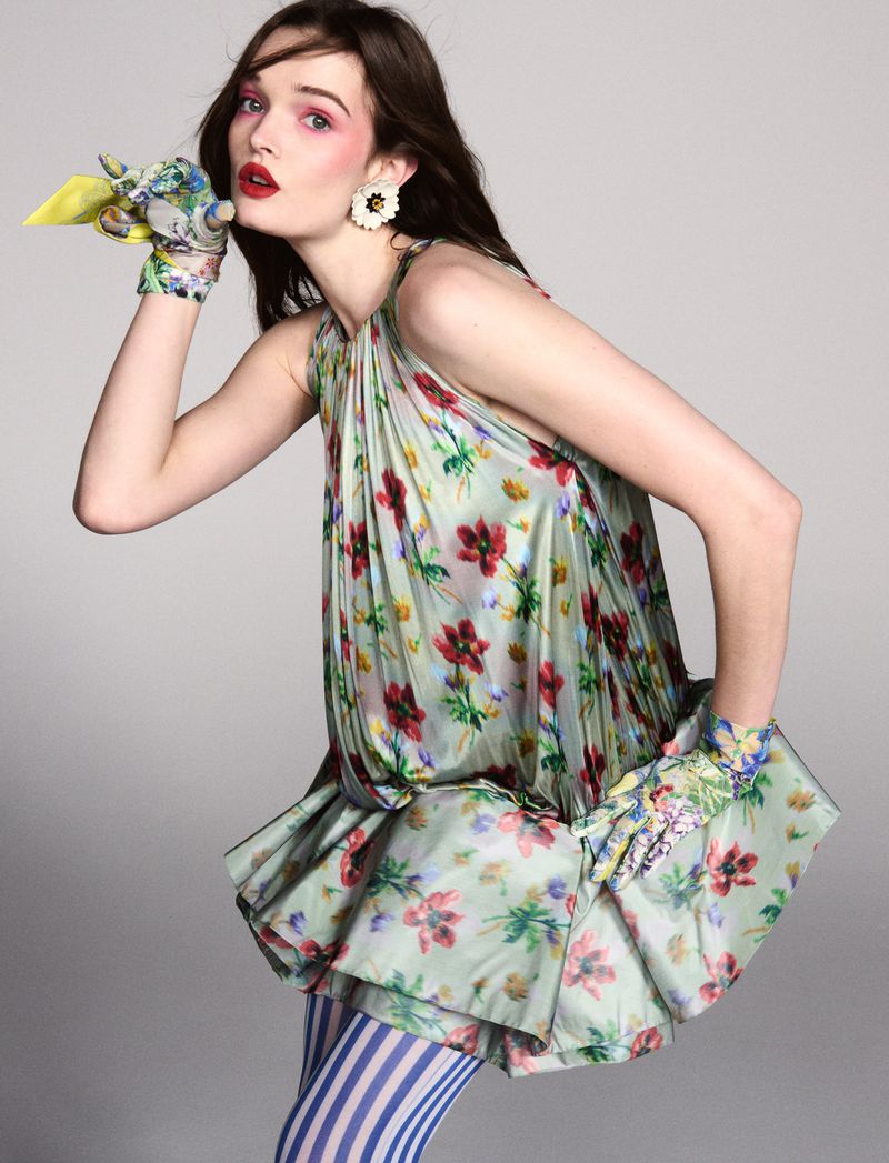 Elegant woman in floral dress and gloves, striped leggings, poses in studio with a playful expression, creating a whimsical fashion statement.