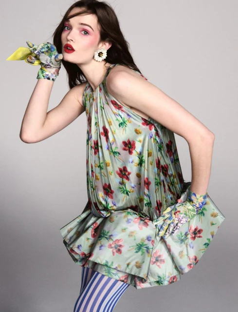 Elegant woman in floral dress and gloves, striped leggings, poses in studio with a playful expression, creating a whimsical fashion statement.