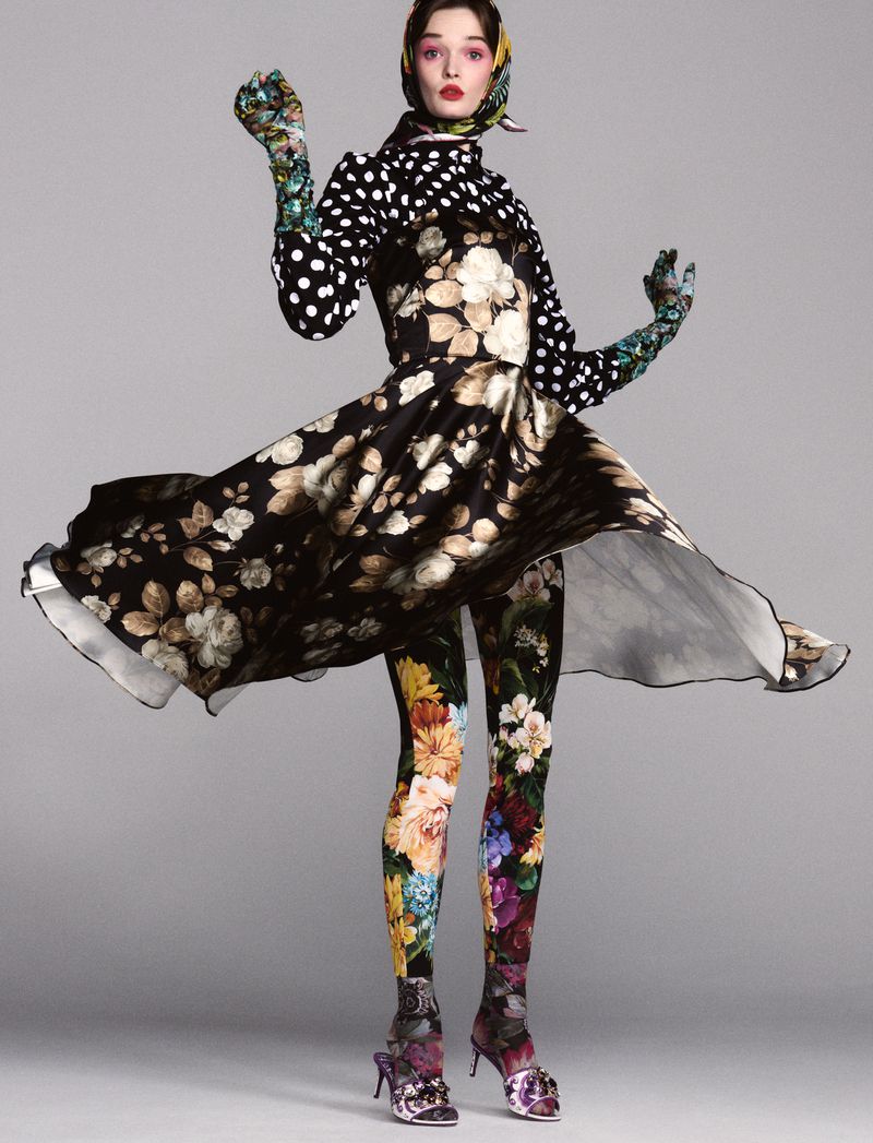 A fashion model in a floral dress and leggings poses against a grey backdrop, accented with polka dots and a scarf, showcasing a unique style.