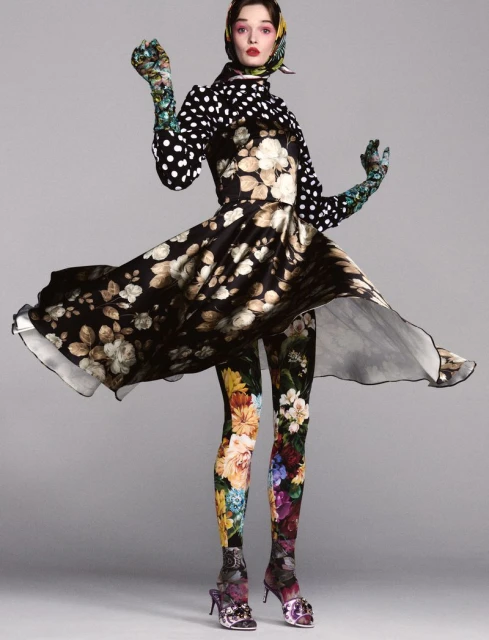 A fashion model in a floral dress and leggings poses against a grey backdrop, accented with polka dots and a scarf, showcasing a unique style.