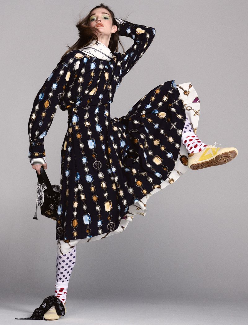 Fashion model strikes a pose in a black long-sleeved dress with chain-link design, paired with polka dot leggings and sneakers against a gray backdrop.