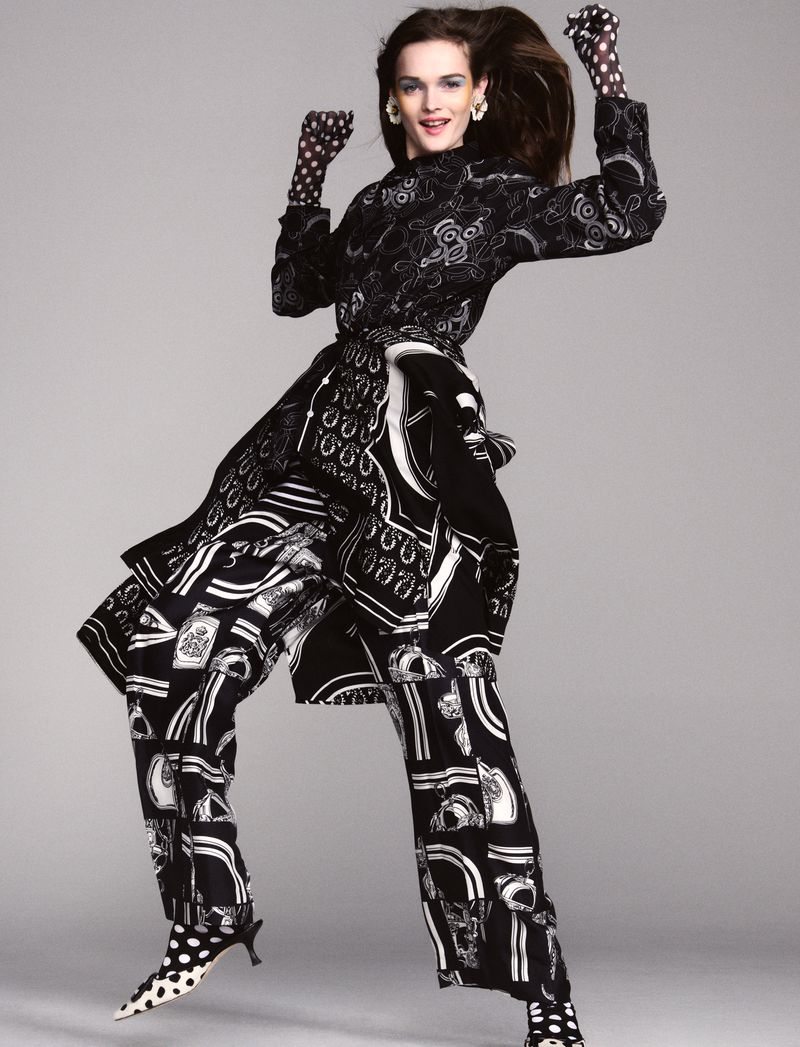 A fashion model jumps in the air, wearing a patterned black and white outfit, gloves, and shoes against a solid grey backdrop.