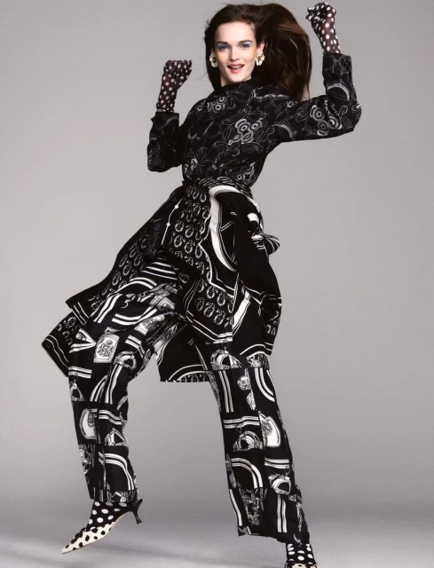 A fashion model jumps in the air, wearing a patterned black and white outfit, gloves, and shoes against a solid grey backdrop.
