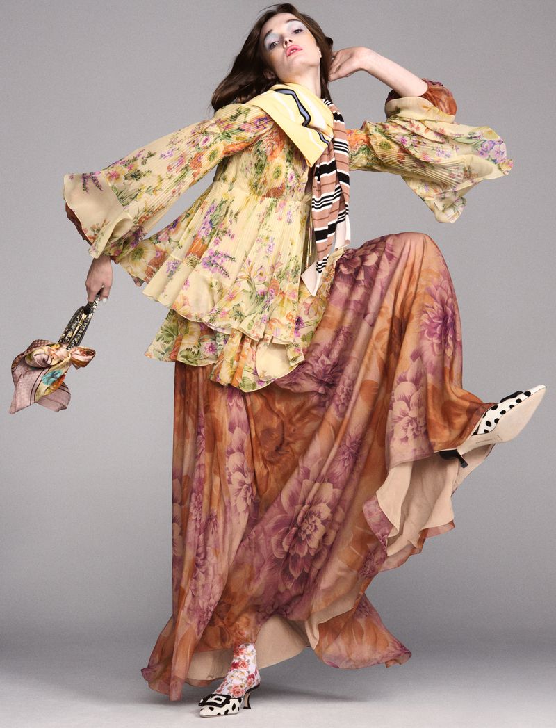 A woman strikes a dynamic pose in a floral-patterned dress with a tiered top and a long skirt, accentuated by patterned accessories.