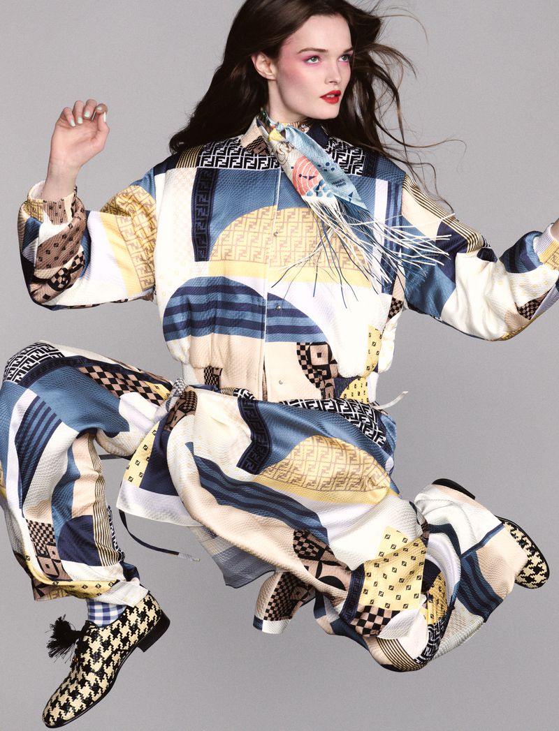 Fashion shot of a woman in patterned Fendi jacket and pants, captured in mid-air, styled with a silk scarf and patterned shoes in a grey studio.