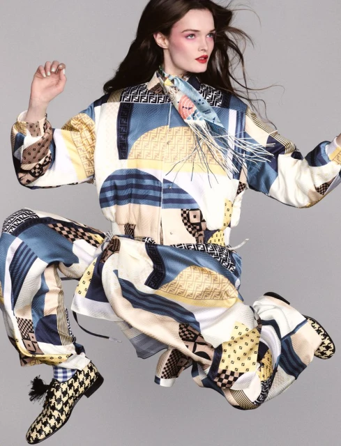 Fashion shot of a woman in patterned Fendi jacket and pants, captured in mid-air, styled with a silk scarf and patterned shoes in a grey studio.
