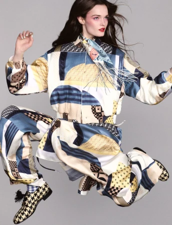 Fashion shot of a woman in patterned Fendi jacket and pants, captured in mid-air, styled with a silk scarf and patterned shoes in a grey studio.