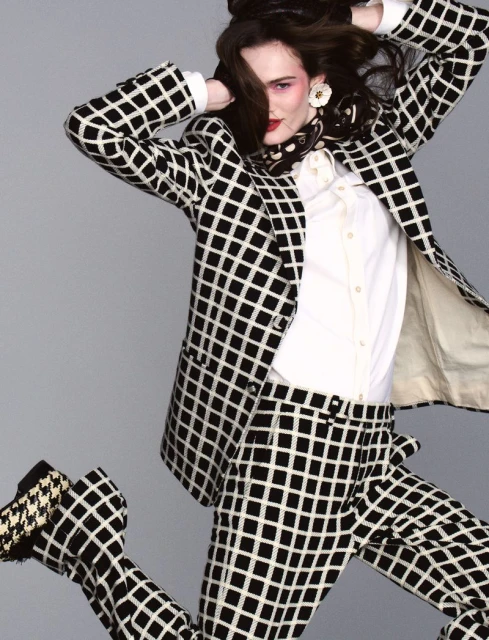 A person with dark hair poses in a black and white checked suit, white collared shirt, and matching heels against a gray backdrop.