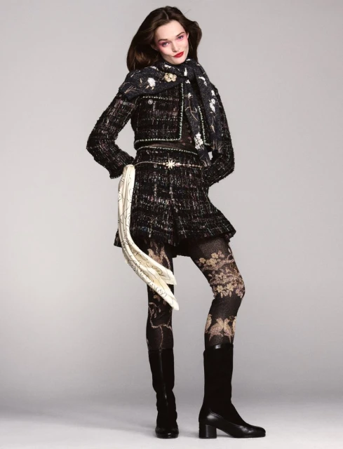 Fashion model wears patterned jacket, skirt, scarf, and tights in studio. She poses against a neutral backdrop, looking at the camera.