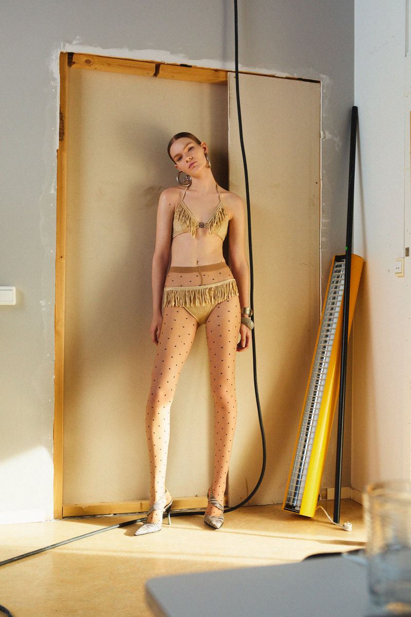 A young, slender woman in a fringed bikini and polka-dot tights poses in a bare room with natural light, showcasing fashion and style.