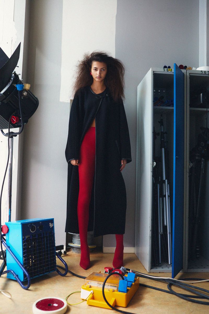 A woman in a black coat and red tights poses in a studio setting with photography equipment, cables, and metal storage cabinets in the background.