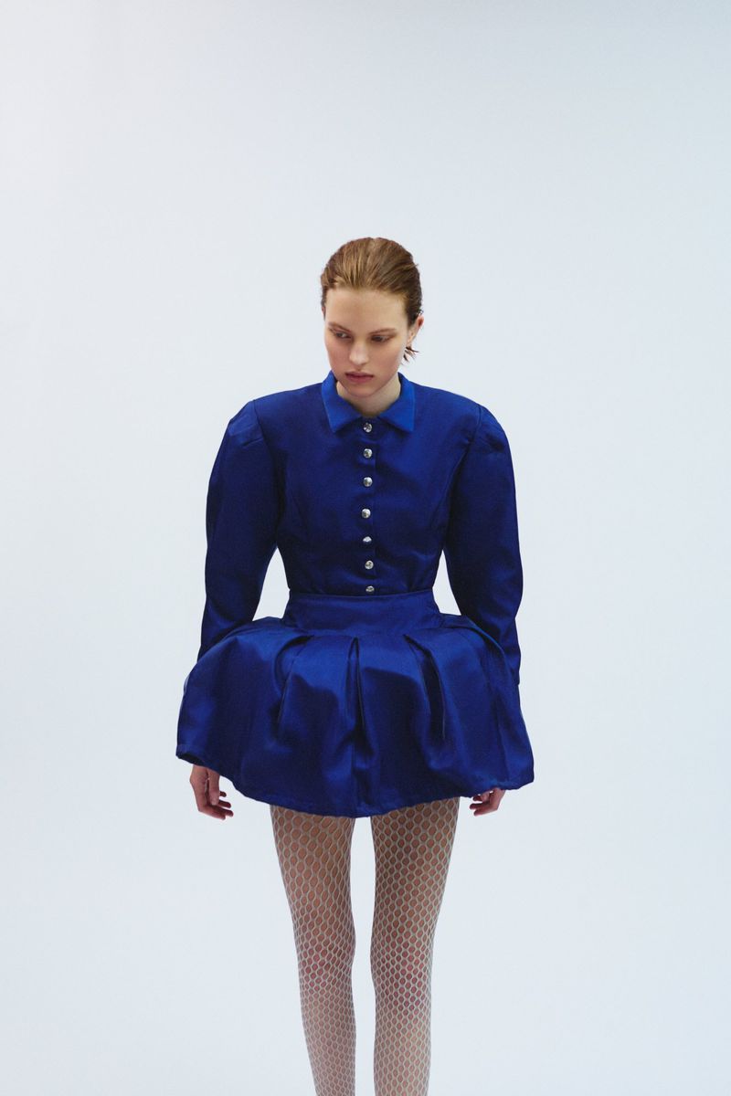 Fashion model in a studio wearing a blue shirtwaist dress with a full skirt and patterned stockings in front of a white background.