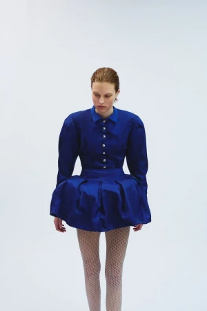 Fashion model in a studio wearing a blue shirtwaist dress with a full skirt and patterned stockings in front of a white background.