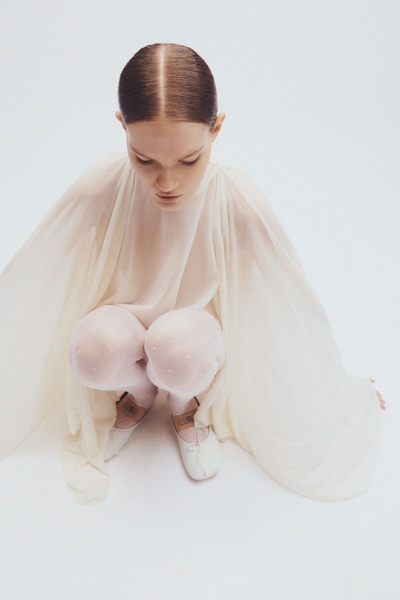 Woman in flowing sheer white dress crouches indoors against a seamless white background, head bowed, ballet shoes on her feet.