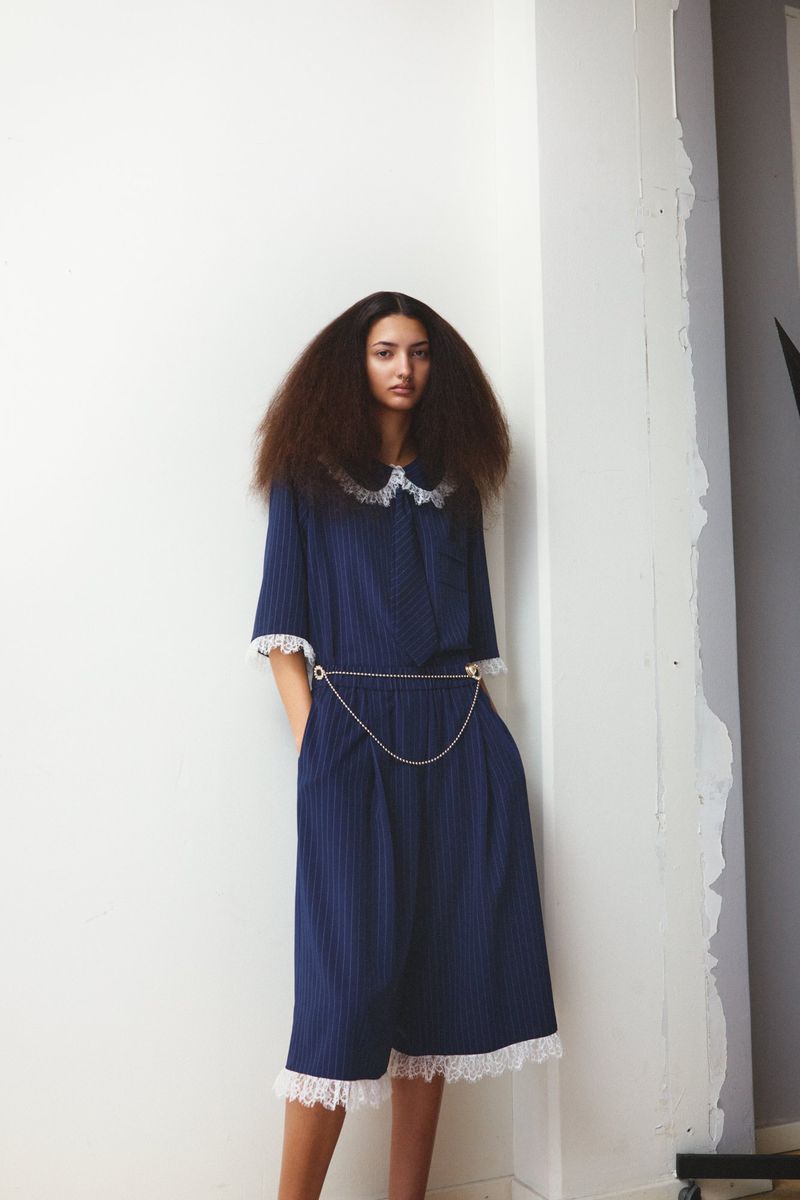 A woman with voluminous brown hair poses indoors wearing a navy striped dress with lace trim and a silver chain belt detail.