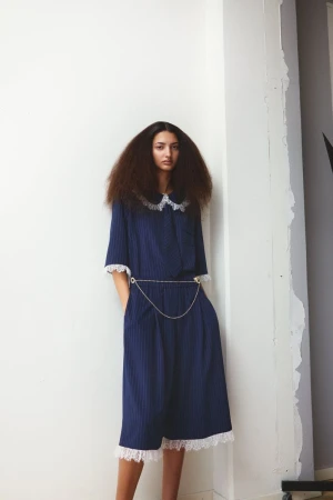 A woman with voluminous brown hair poses indoors wearing a navy striped dress with lace trim and a silver chain belt detail.