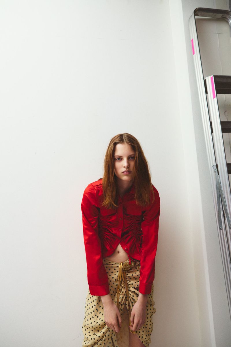 Young woman poses in a red top and patterned skirt indoors near a white wall and stepladder, creating a fashion editorial style portrait.